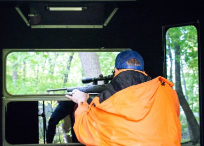 back of hunter aiming firearm through window in hard sided hunting blind